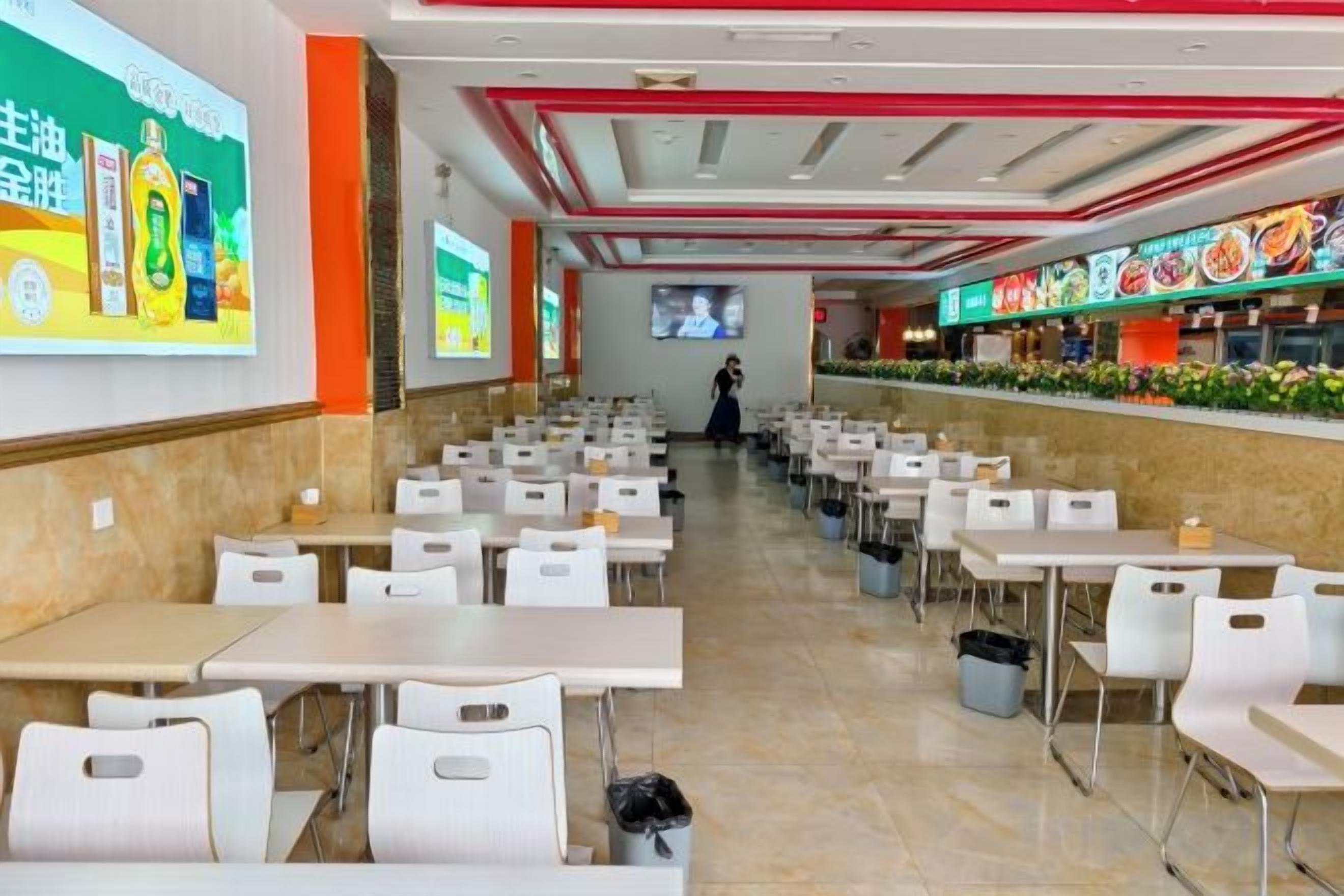 A cafeteria with rows of tables and chairs, a projection screen on the wall, and a food counter with a menu.