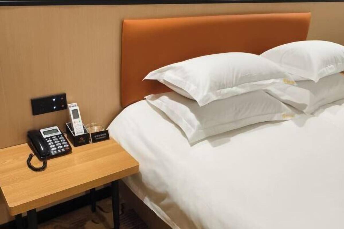 A hotel room with a bed, white pillows, a wooden nightstand with a phone, a glass of water, and a small box.