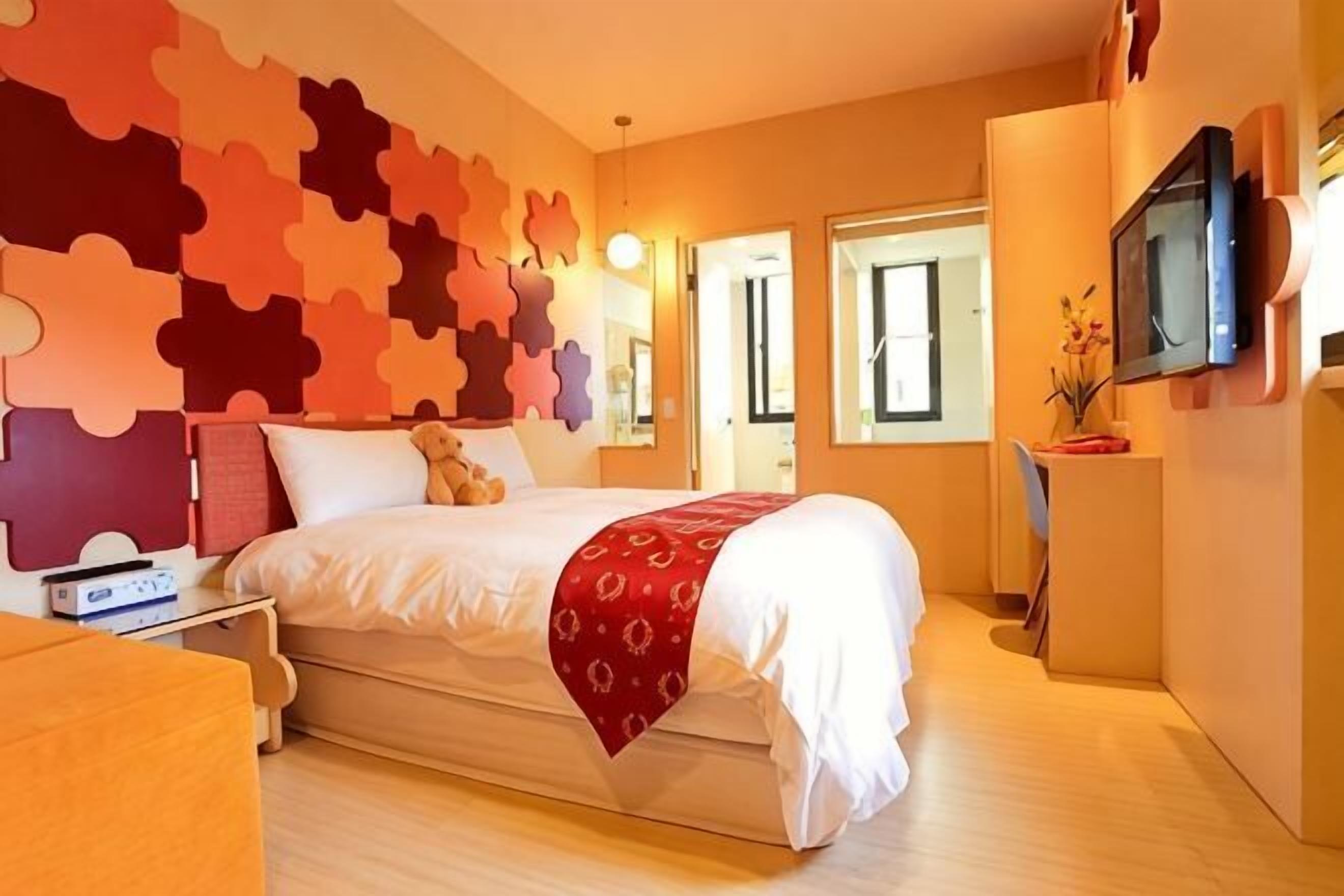 A hotel room with a bed, a TV, a desk, and a wall with puzzle piece decor.