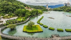 Um parque à beira-rio com uma passarela circular, uma ilha central com uma árvore e edifícios ao fundo.
