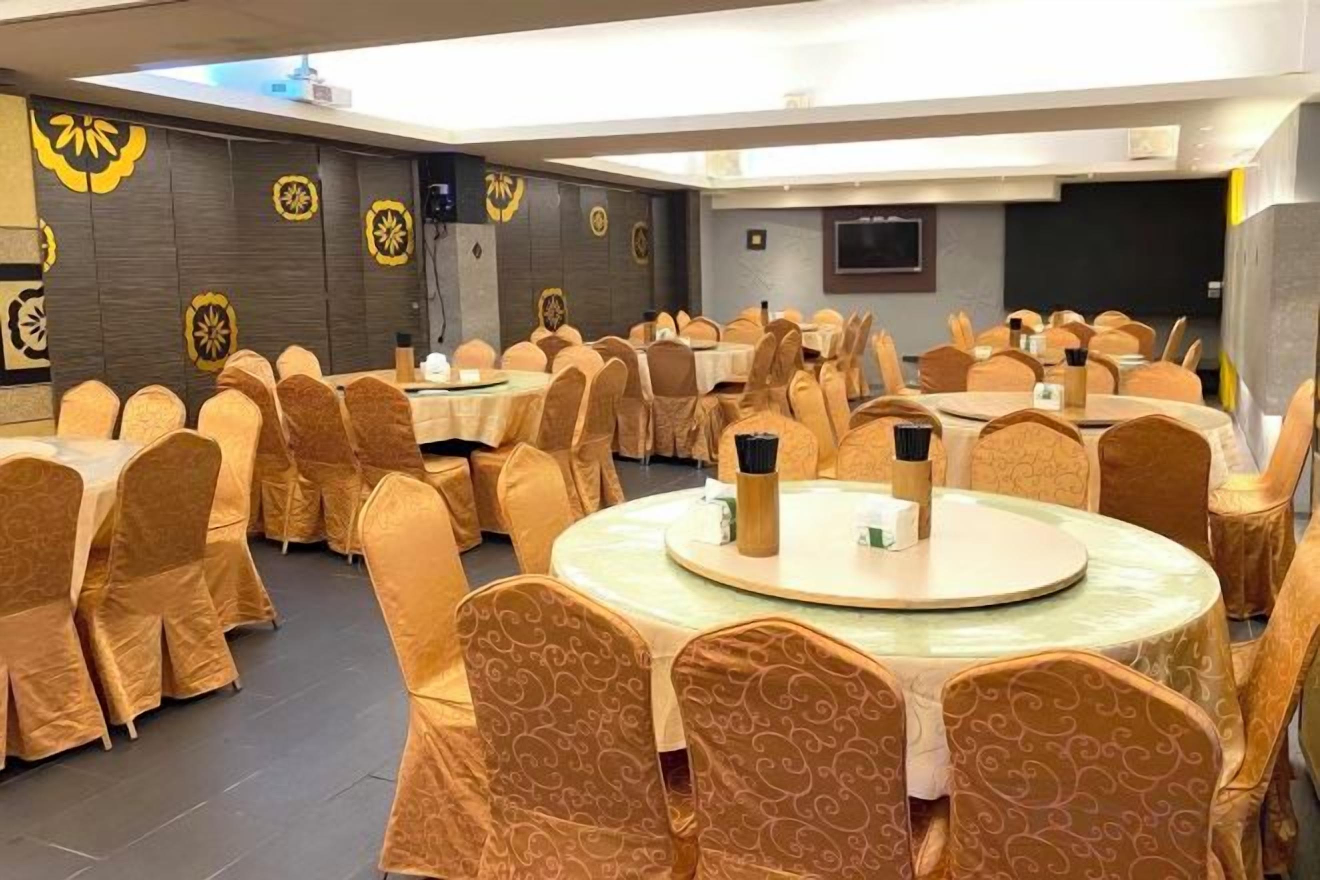 A banquet hall with round tables and chairs, a flat-screen TV, and decorative wall patterns.