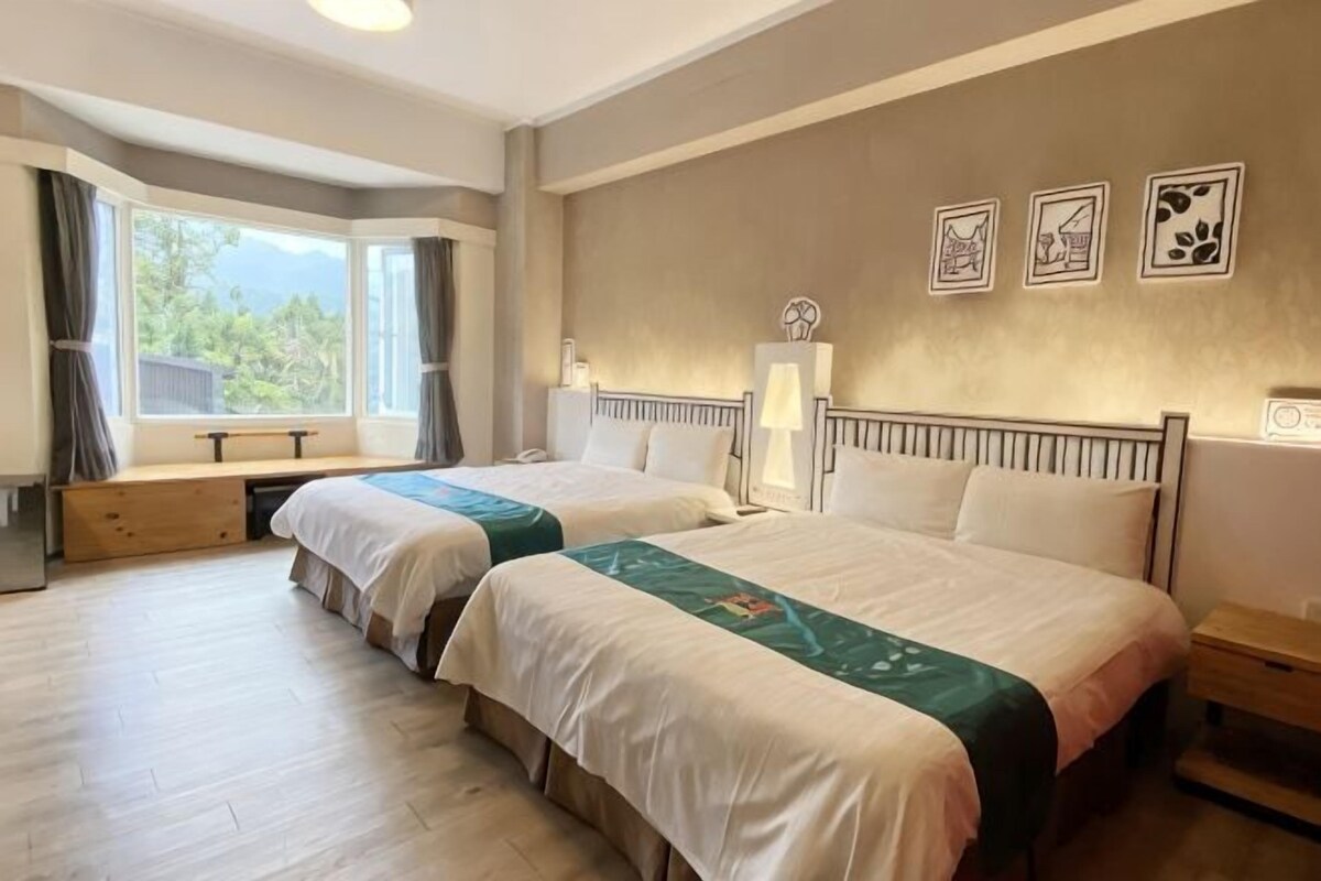 A hotel room with two beds, a window with curtains, a bedside table, and a wall with framed artwork.