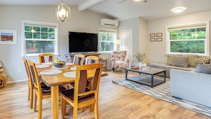 Un salon spacieux avec une table à manger en bois, des sièges confortables et de grandes fenêtres donnant sur un paysage verdoyant.