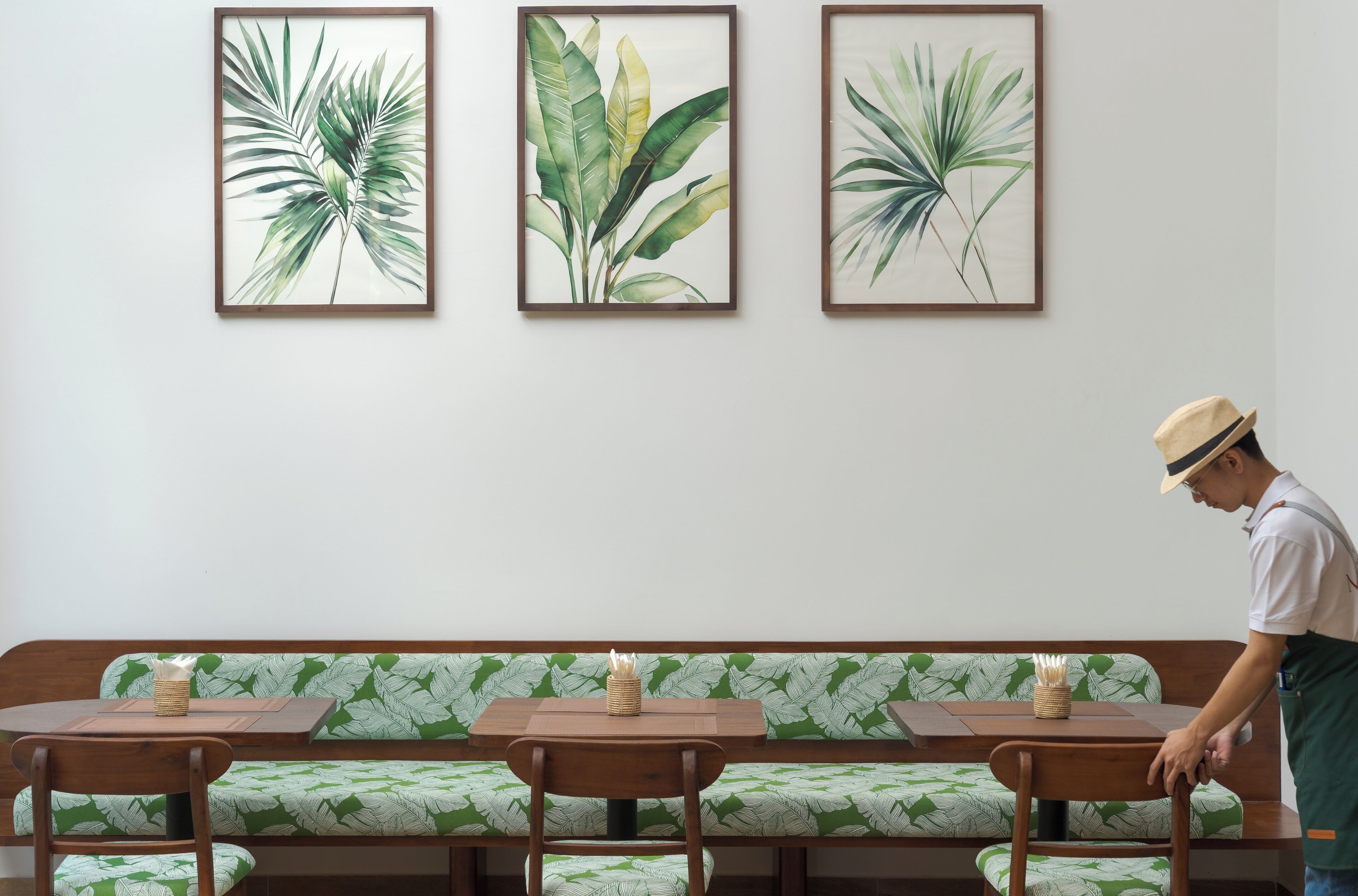 Een eetruimte met een bank en tafels, met botanische prints in de wanden, en een persoon in een schort.