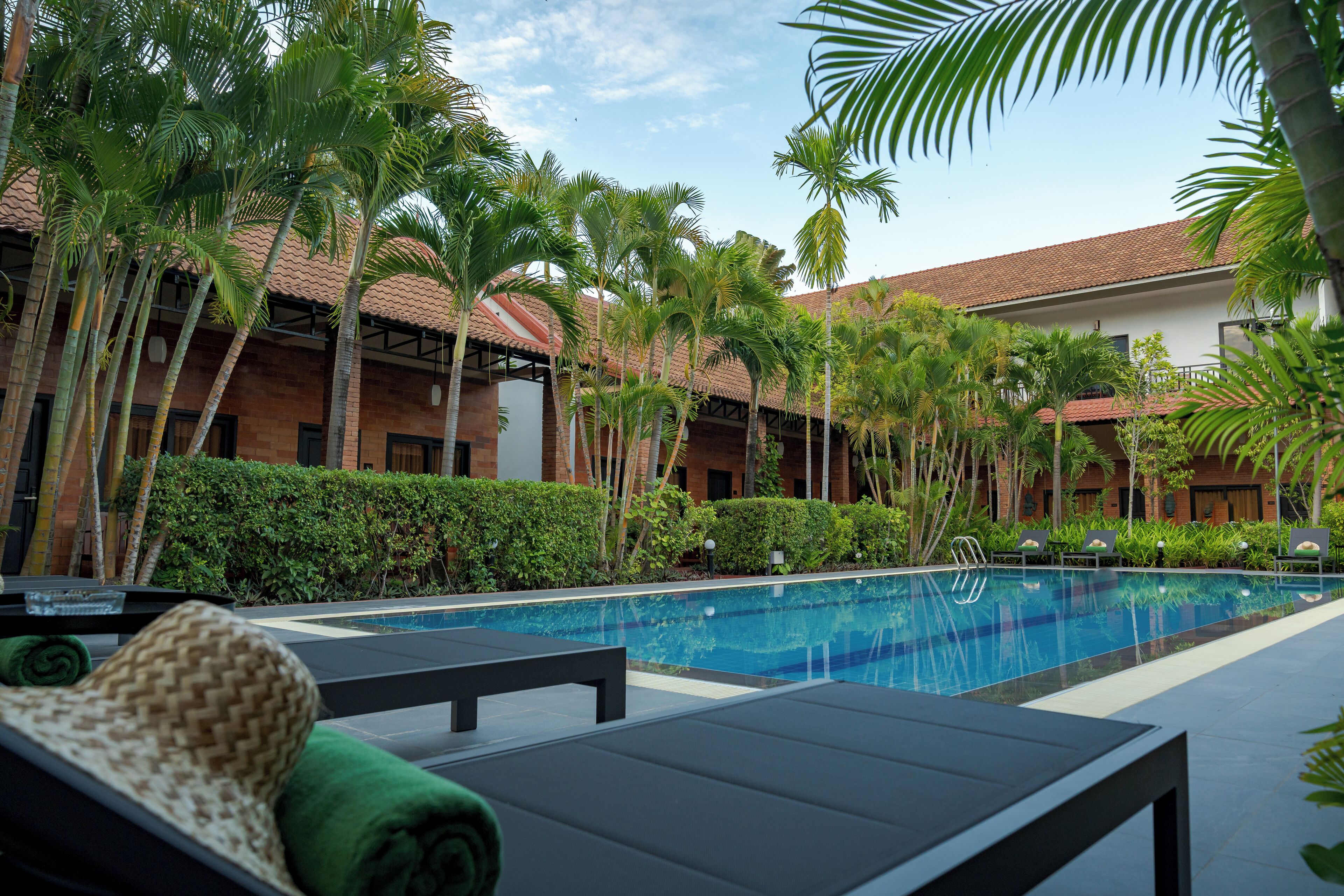 A poolside area with lounge chairs, a swimming pool, palm trees, and a building with a tiled roof.