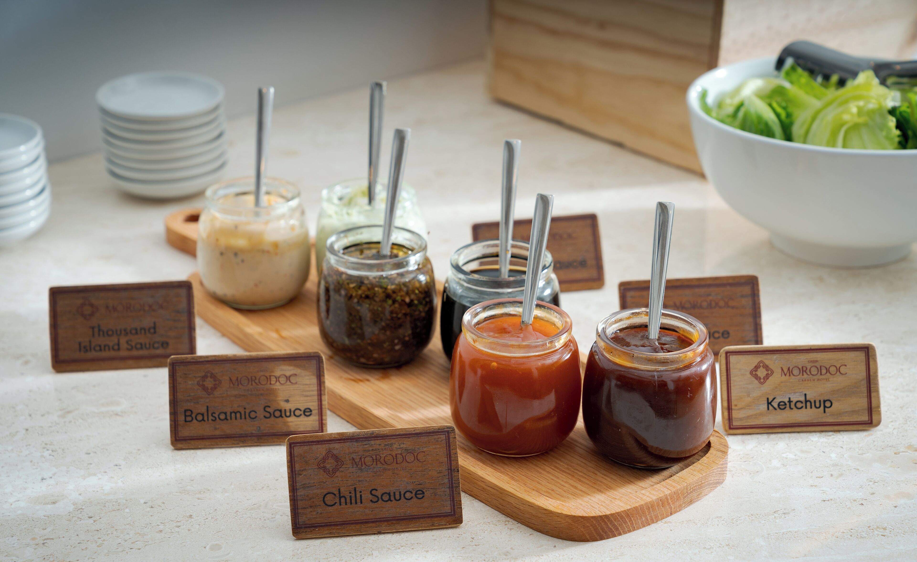 A variety of condiments with labels on a wooden board, including Thousand Island Sauce, Balsamic Sauce, Chili Sauce, and Ketchup.