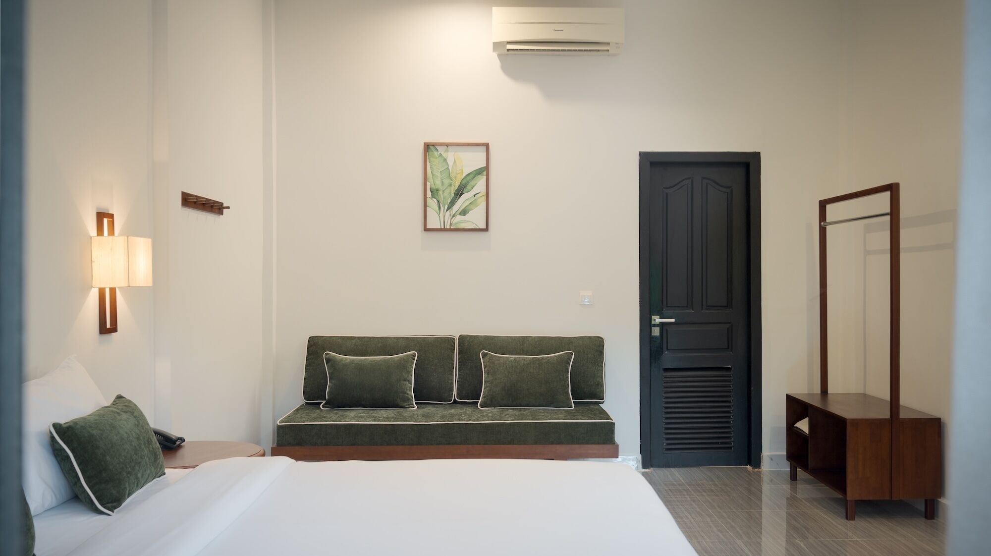Una habitación de hotel con cama, sofá con cojines, armario, espejo, aire acondicionado de pared y una planta en marco.