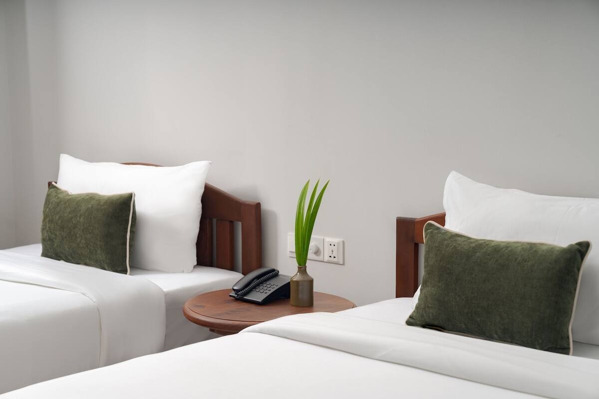 Una camera d'hotel con due letti, ciascuno con biancheria da letto bianca e cuscini verdi. Tra i letti, un piccolo tavolino in legno con un telefono e un vaso con una pianta.