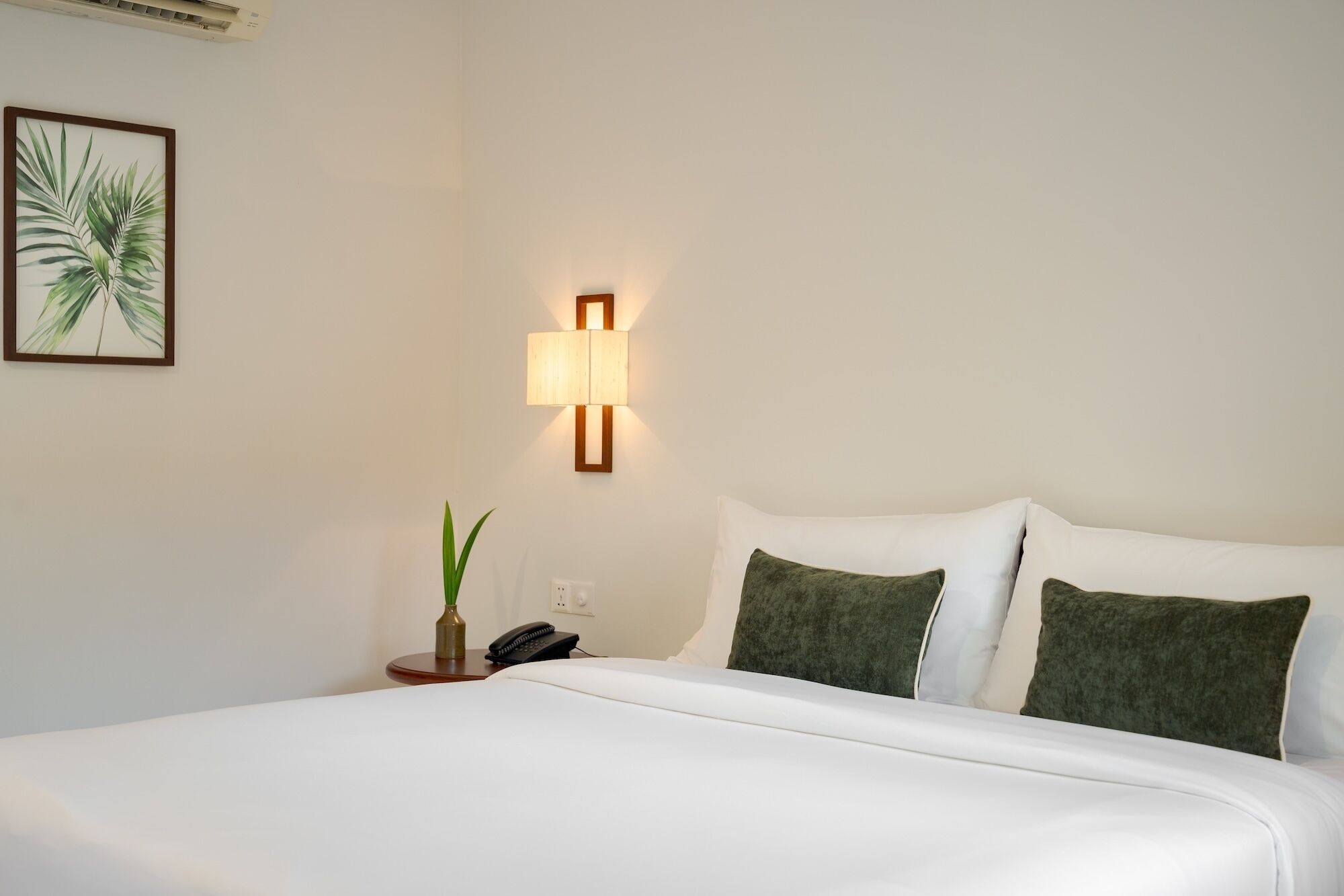 A hotel room with a bed, a wall-mounted lamp, a plant, and a phone on a bedside table.