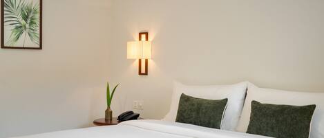A hotel room with a bed, a wall-mounted lamp, a plant, and a phone on a bedside table.