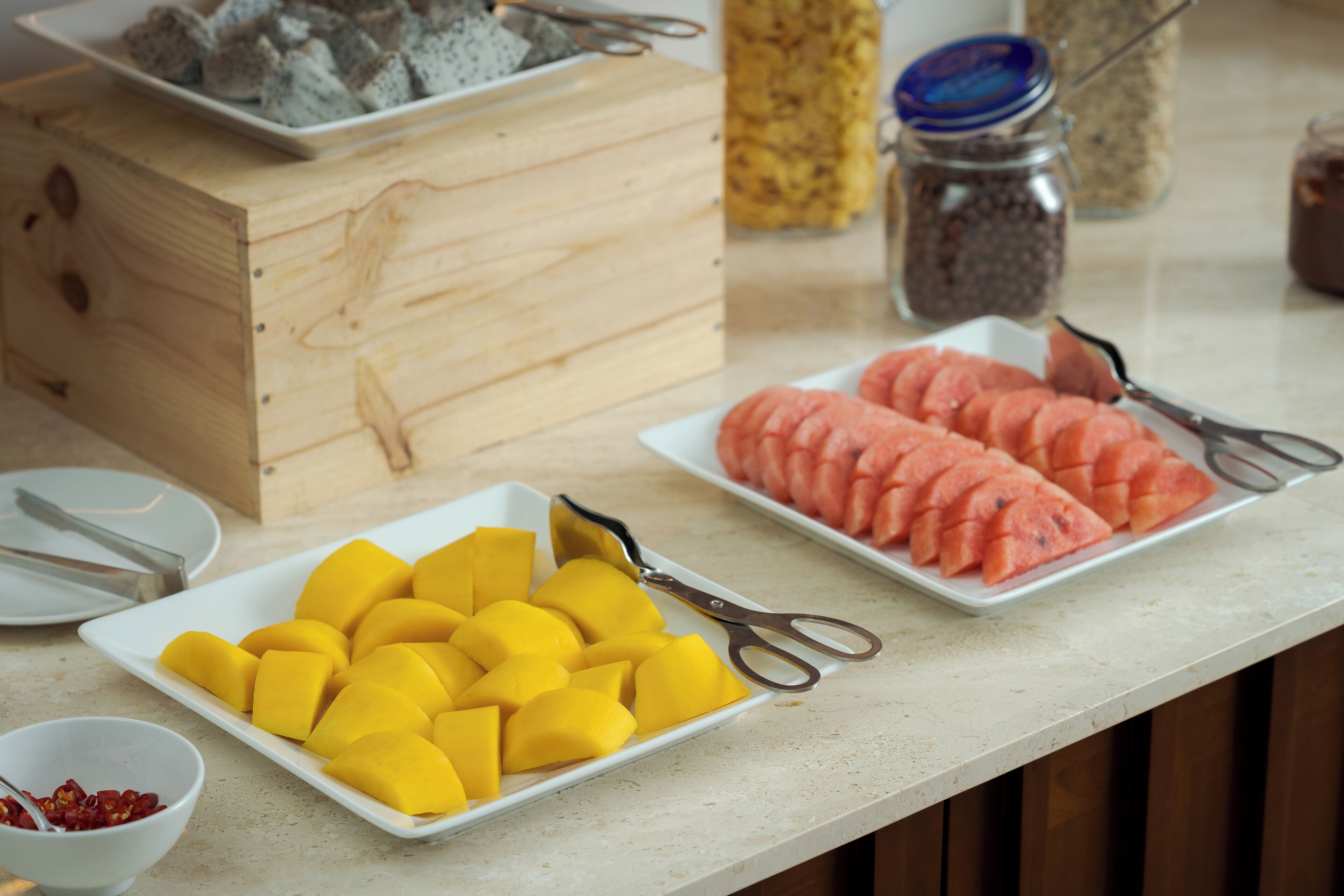 A buffet table with plates of sliced mango and watermelon, a wooden box, and a jar of spices.