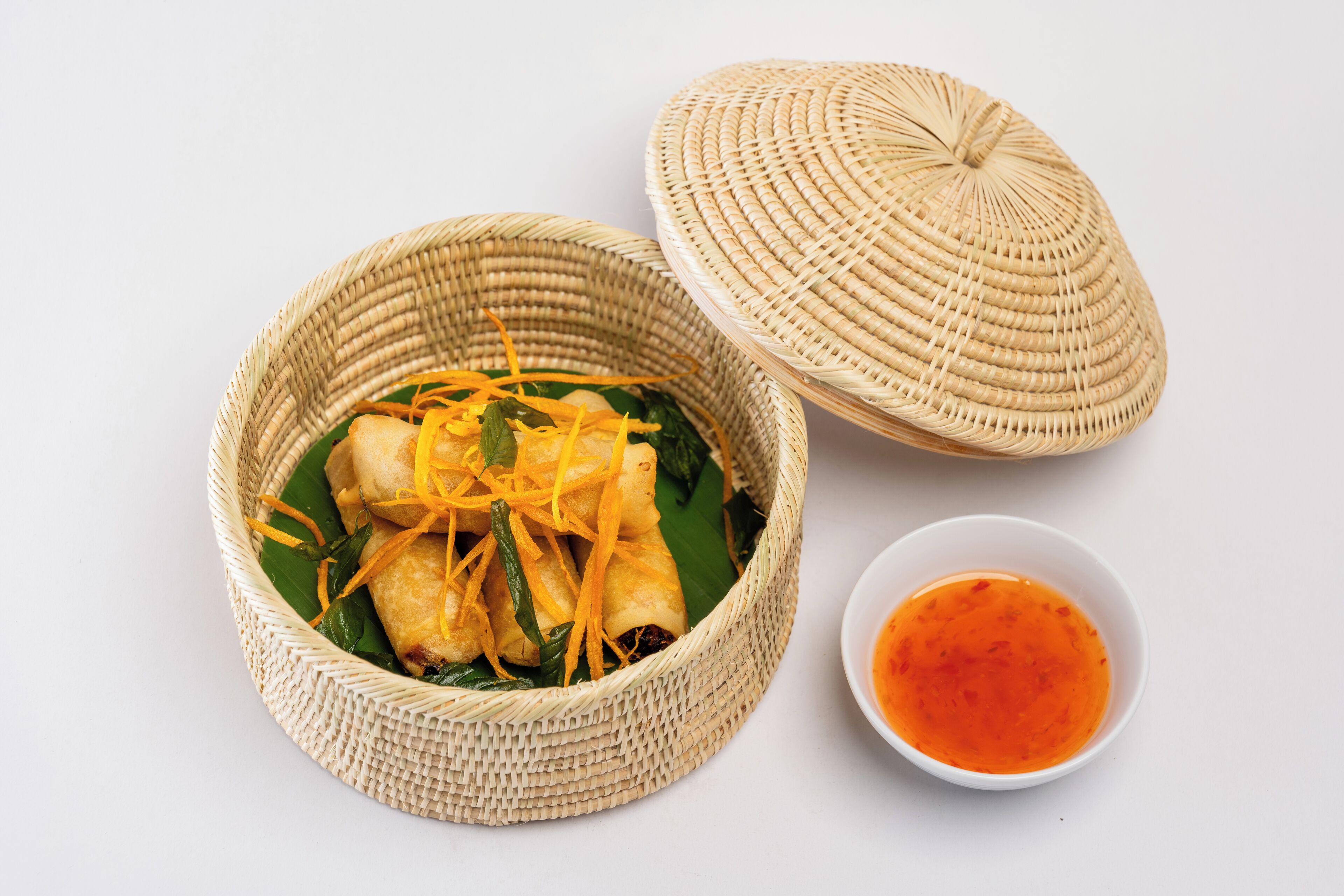 A woven basket with food, a lid, and a small bowl of sauce.