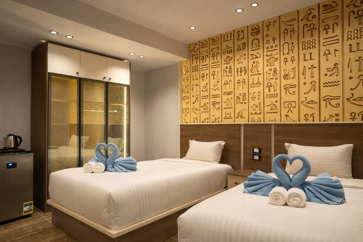 A hotel room with two beds, a minibar, and a wall decorated with ancient Egyptian hieroglyphs.