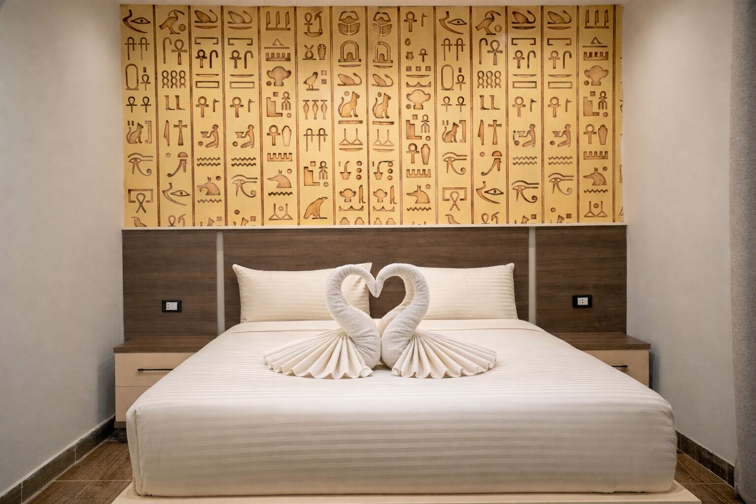 Ein Hotelzimmer mit einem Bett, das mit einer schwanenförmigen Handtuchdeko verziert ist, einer Kopfteil mit ägyptischen Wandmalereien und einem Nachttisch aus Holz.