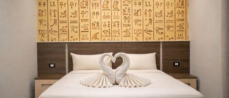 Ein Hotelzimmer mit einem Bett, das mit einer schwanenförmigen Handtuchdeko verziert ist, einer Kopfteil mit ägyptischen Wandmalereien und einem Nachttisch aus Holz.