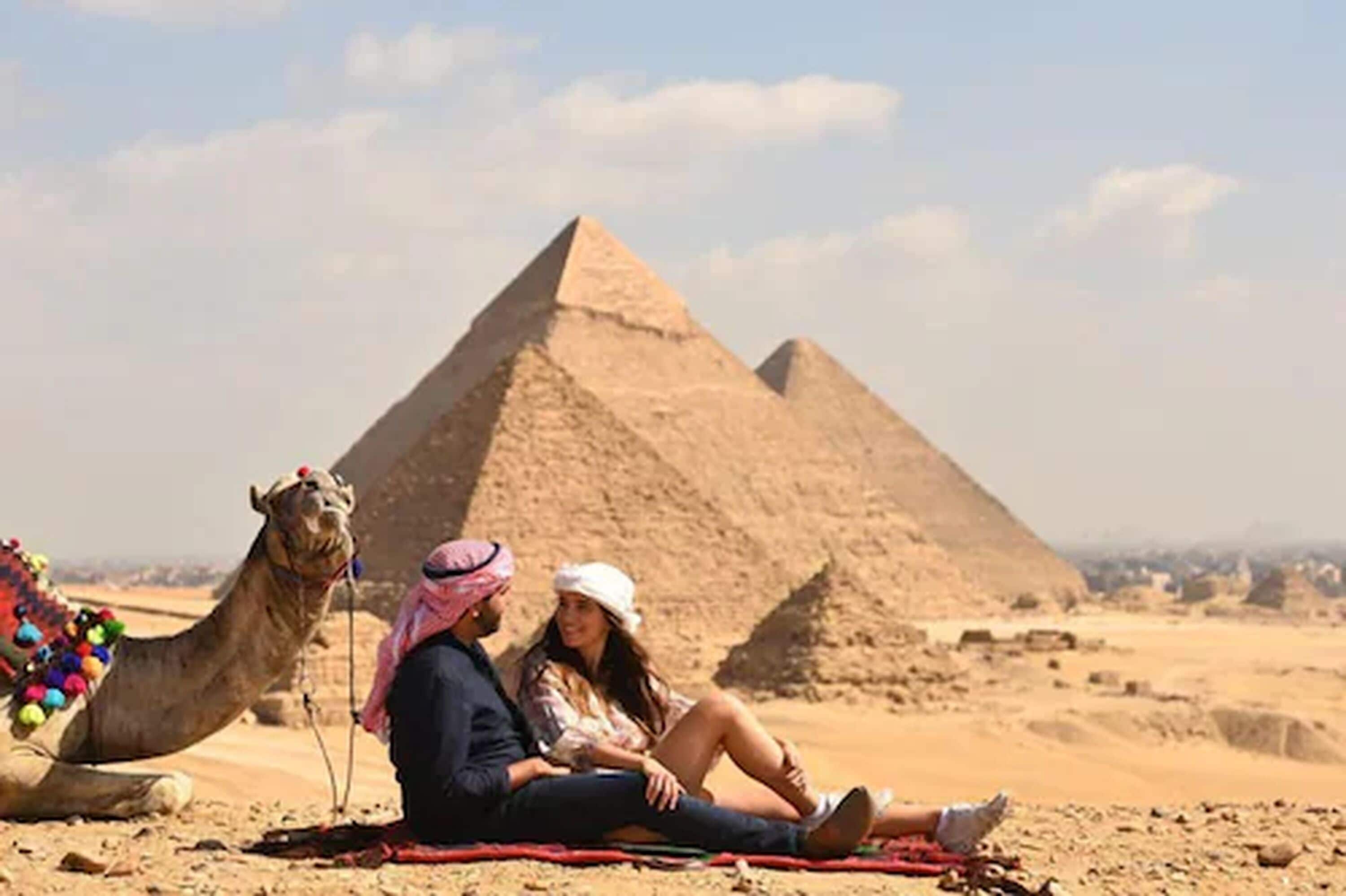 Ein Paar sitzt auf einer Decke, vor ihnen ein Kamel vor der Pyramide von Gizeh.