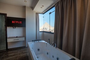 A modern bathroom with a large bathtub, a wall-mounted television displaying 'NETFLIX', and a view of a pyramid through a window.