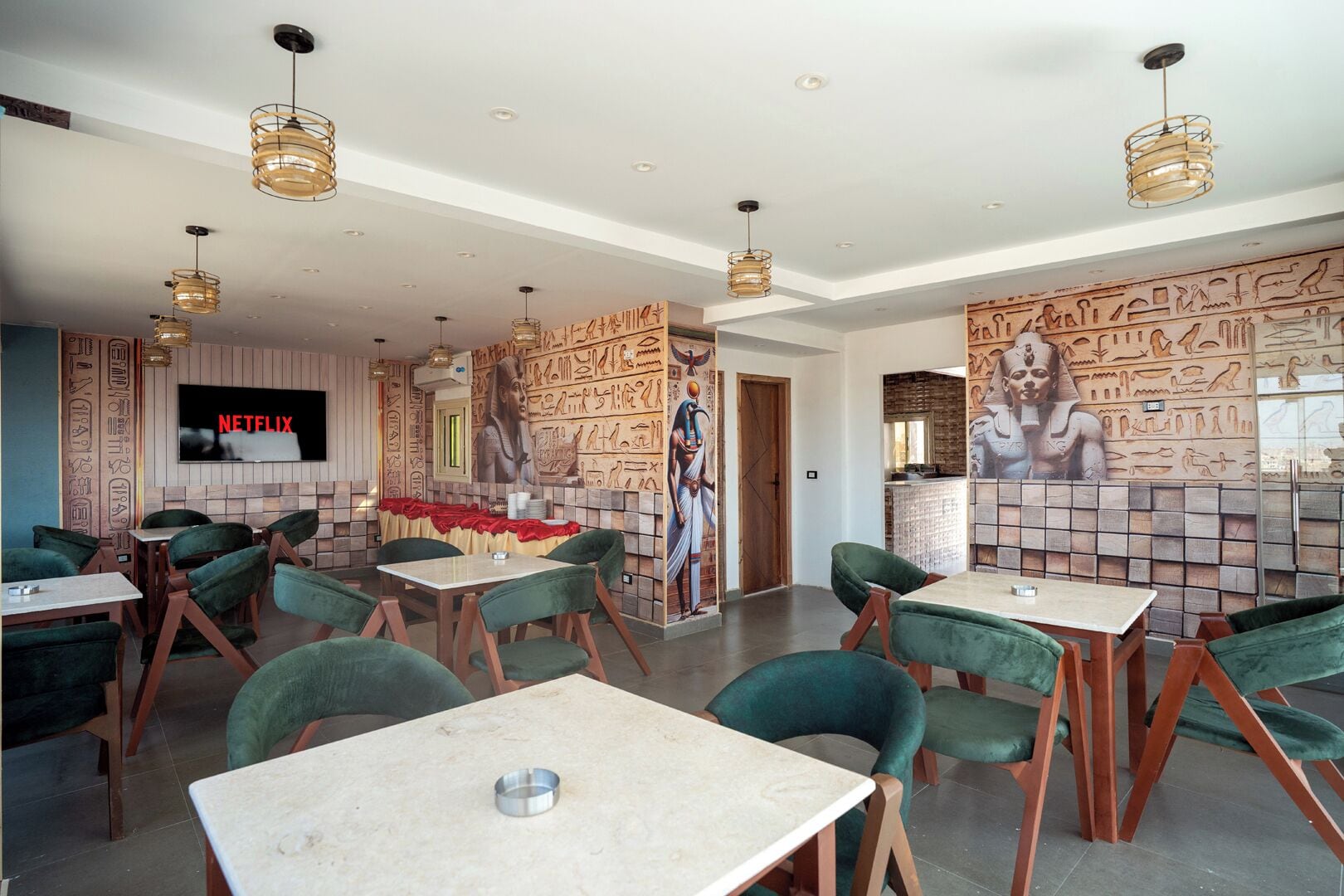 Un restaurant avec des chaises à revêtement vert, des tables en marbre et des peintures murales représentant l’art de l’Égypte ancienne.