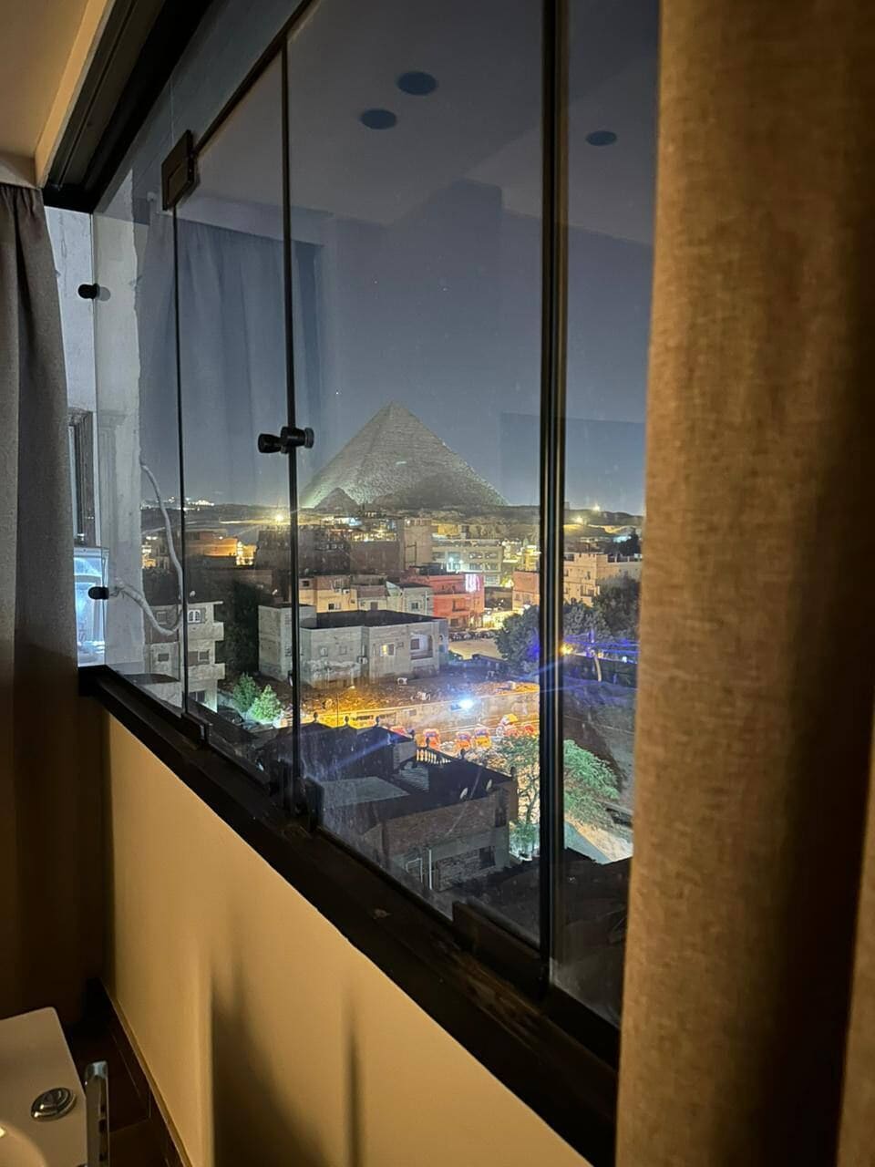Une pièce dotée d’une grande fenêtre donnant sur une vue panoramique de la ville la nuit, avec des bâtiments illuminés et une montagne en arrière-plan.