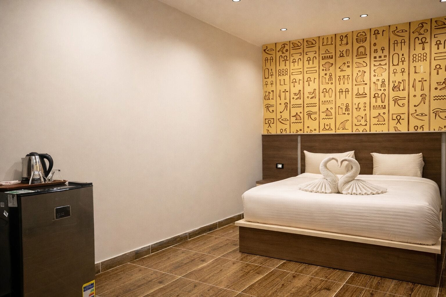 A hotel room with a bed, a wooden headboard, a wall with hieroglyphs, and a minibar.