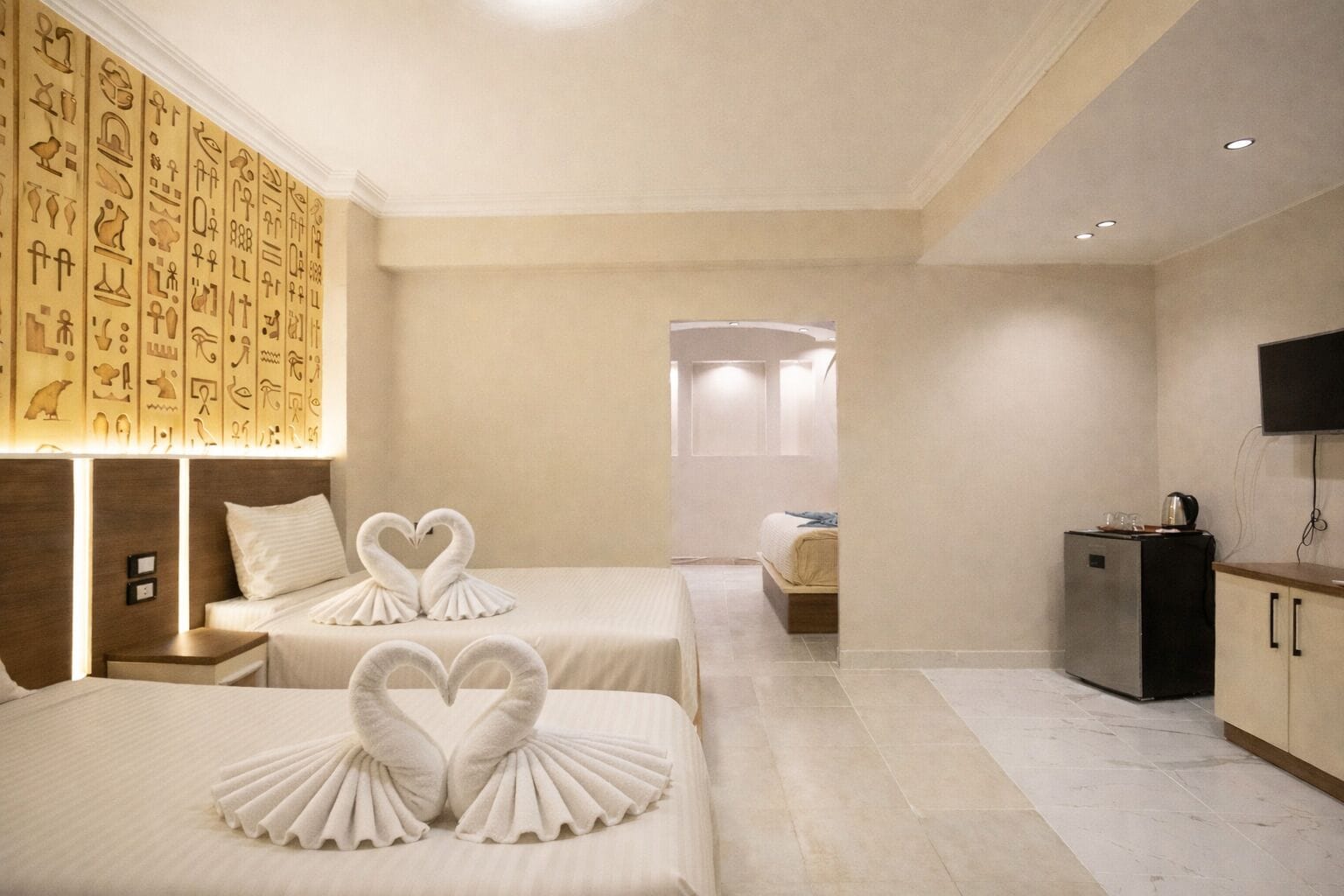 A hotel room with two beds, a minibar, a television, and a wall with hieroglyphs.
