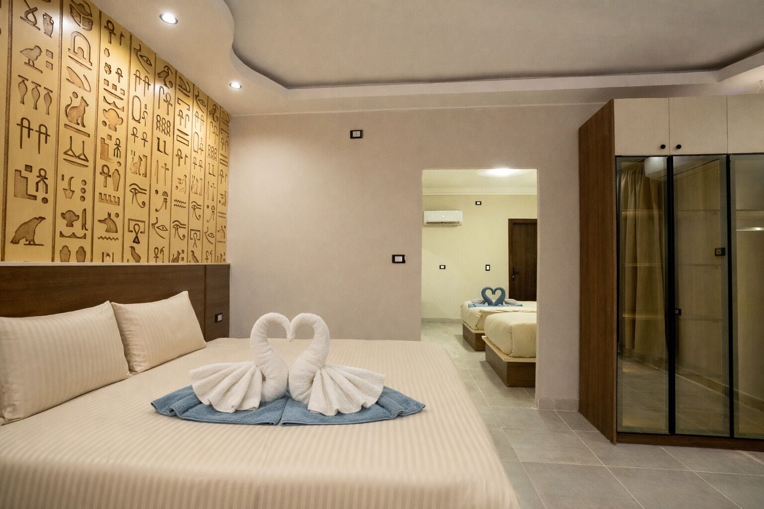 Quarto de hotel com cama, parede de hieróglifos, guarda-roupa e banheiro visível através de uma porta aberta.