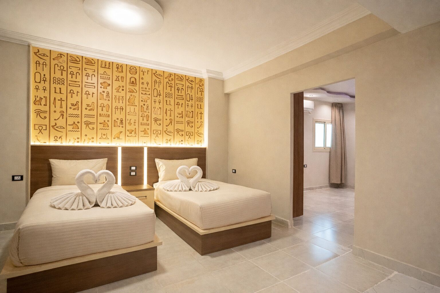 Habitación de hotel con dos camas, cada una adornada con una toalla en forma de corazón, una pared con jeroglíficos y un diseño moderno.