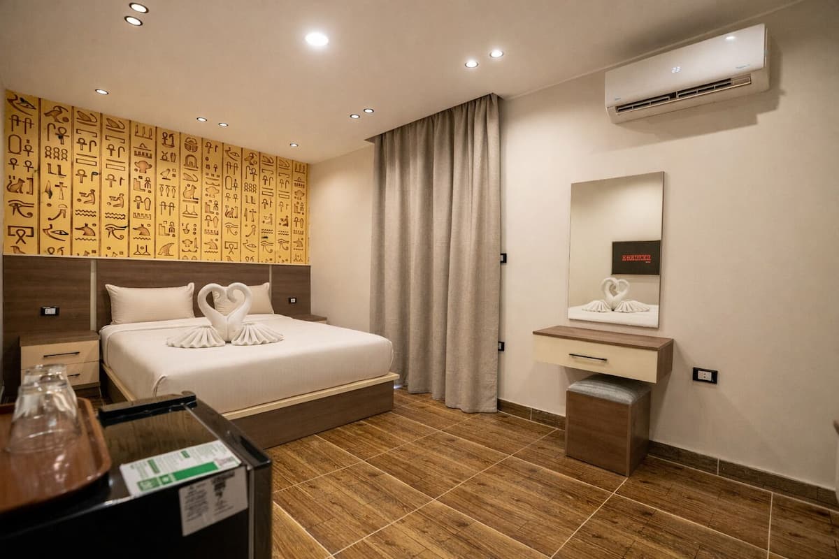 A hotel room with a bed, a wooden headboard, a wall with hieroglyphs, a mirror, a nightstand, and a wooden floor.