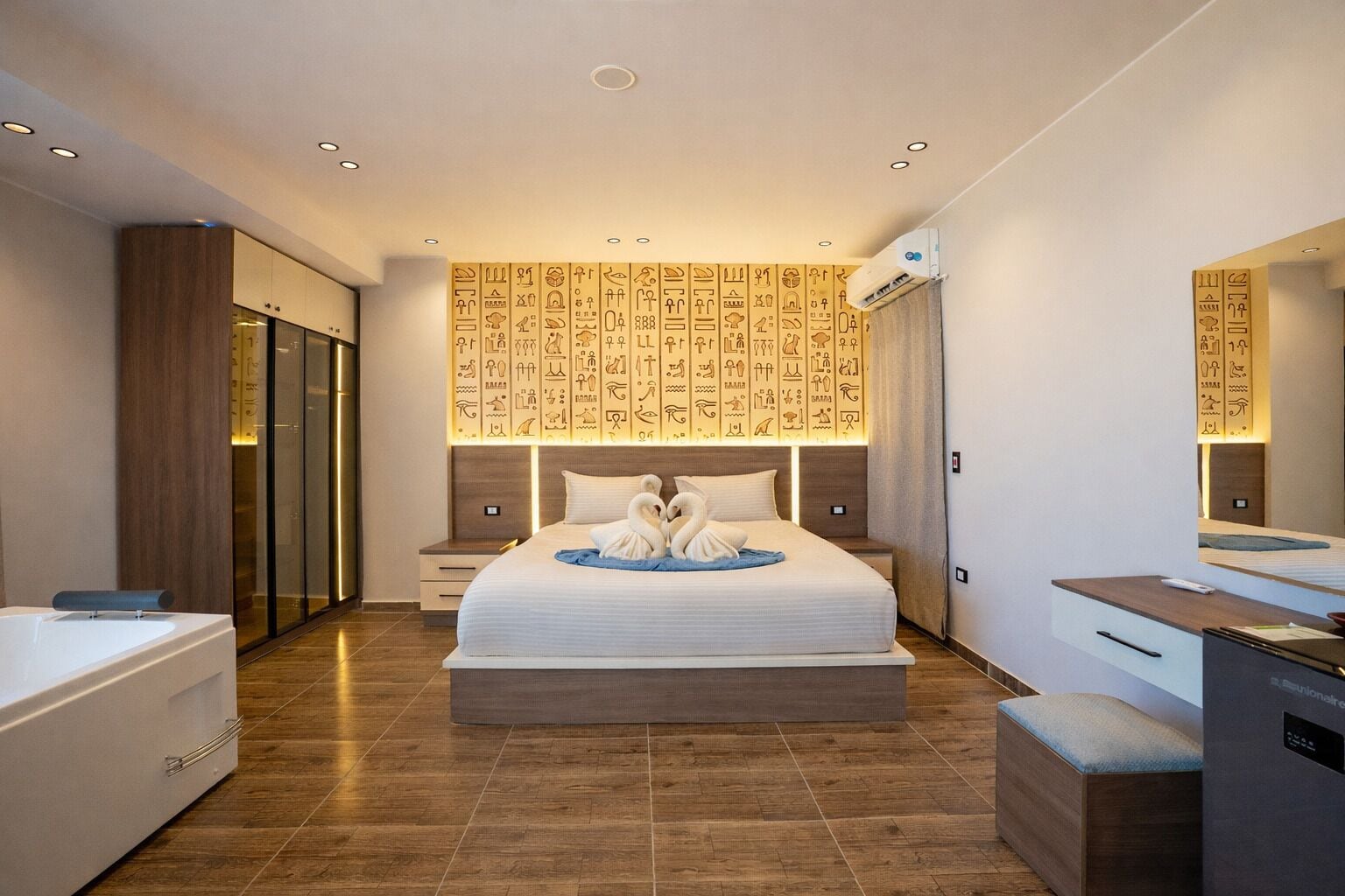 Una habitación de hotel moderna con una cama grande, un armario de madera, un baño con bañera y una pared con jeroglíficos.