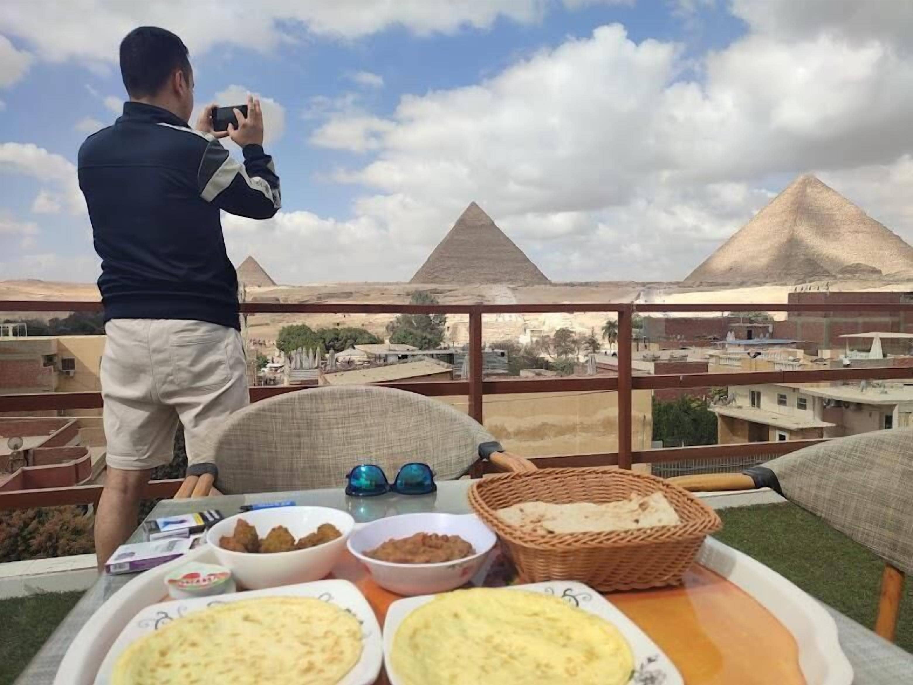A person taking a photo of the pyramids from a balcony with a table of food and drinks.