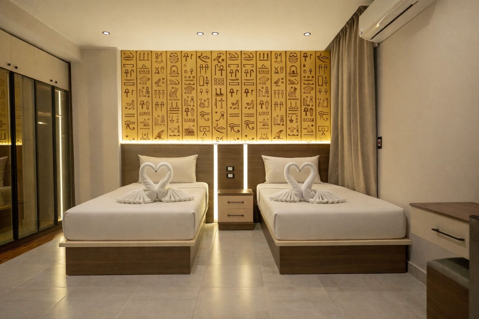 Een hotelkamer met twee bedden, elk met een handdoekarrangement in de vorm van een hart, een muur met Egyptische hiërogliefen, en een modern design.