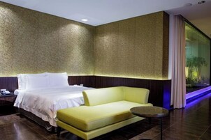 Habitación de hotel con una cama, un sofá amarillo, una mesita redonda y un amplio ventanal con cortinas.