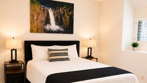 Een slaapkamer met een bed, twee nachtkastjes, een raam met jaloezieën, een aan de muur hangend kunstwerk en een vloerbedekking.