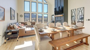 Uma sala de estar ampla, com uma grande mesa de jantar de madeira, assentos confortáveis e vista para o exterior através de amplas janelas.