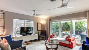 Een ruime woonkamer met een grote flatscreen-televisie, comfortabele zithoeken, een plafondventilator en uitzicht op een tuin door grote ramen.