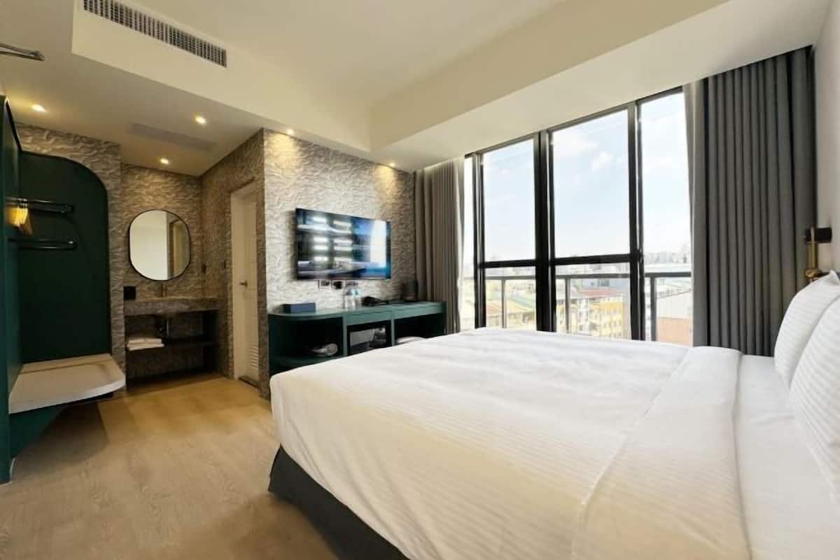 A modern hotel room with a large bed, a TV, a vanity with a mirror, and a view of the city through large windows.