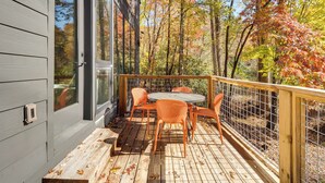 Terraza de madera con una mesa y sillas, rodeada por una barandilla de madera y vistas a árboles otoñales.