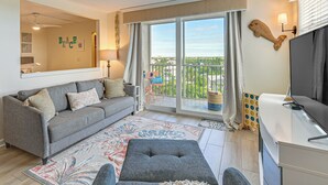 A living room with a grey sofa, a grey ottoman, a large window with a view, and a flat-screen TV on a white table. - Stroll to Bonita Beach Park: Ocean-View Getaway (Bonita Springs)