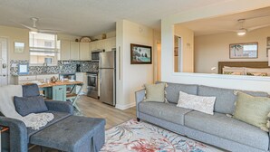 A modern living room with a grey sofa, a grey armchair, and a coffee table. There is a kitchen area with white cabinets and a refrigerator. - Stroll to Bonita Beach Park: Ocean-View Getaway (Bonita Springs)