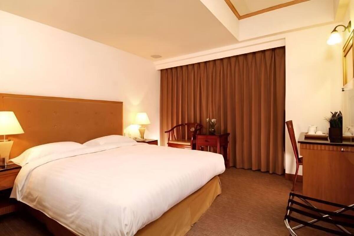 Habitación de hotel con una cama grande, dos mesitas de noche con lámparas, un escritorio, una silla y un ventanal con cortinas.