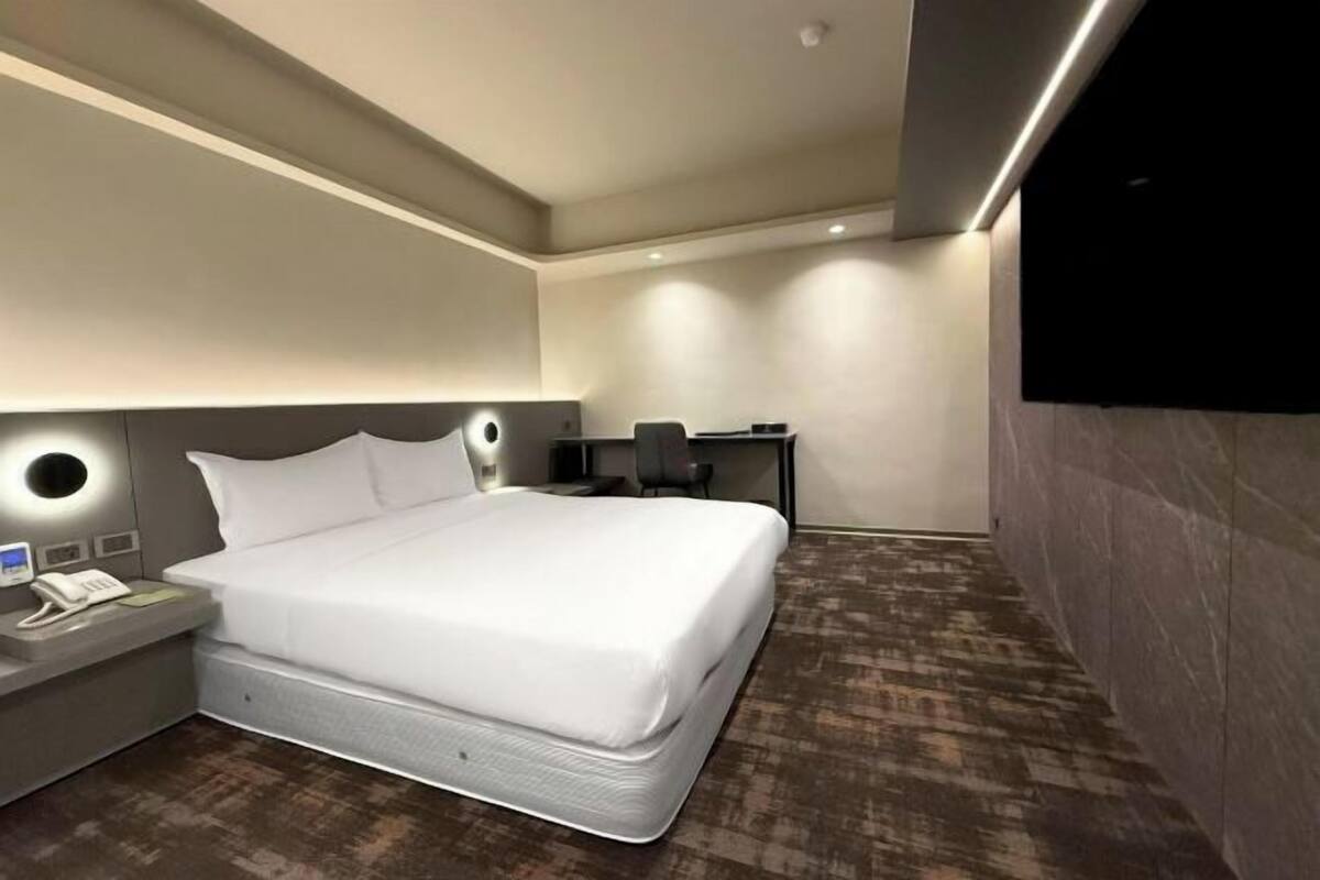 Una habitación de hotel moderna con una cama grande, mesita de noche con teléfono, escritorio con silla y televisor de pantalla plana montado en la pared.