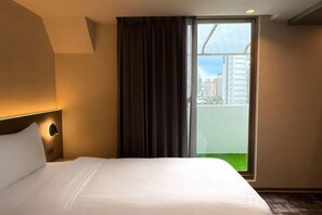 Hotellihuone, jossa on sänky, yöpöytä, ikkuna verhoilla ja näkymä ulkopuolella oleviin rakennuksiin.