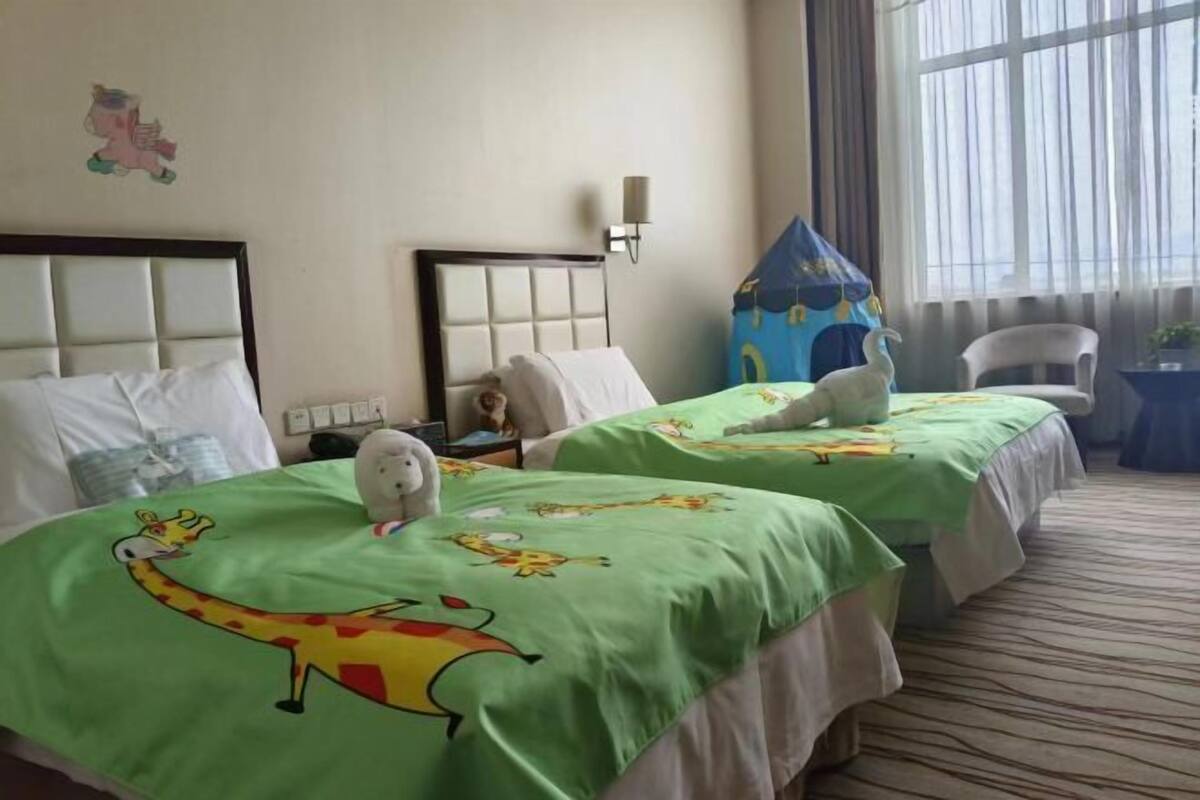 A hotel room with two beds, each with a green bedspread featuring cartoon characters, a white wall with a cartoon character decoration, a nightstand with a lamp, and a window with curtains.