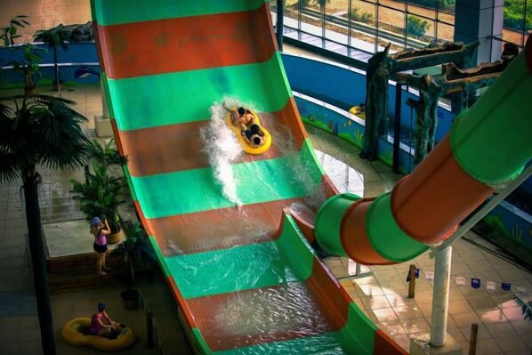 Tobogán de agua con una persona en la parte superior, zona de piscina interior y puesto de socorrista.