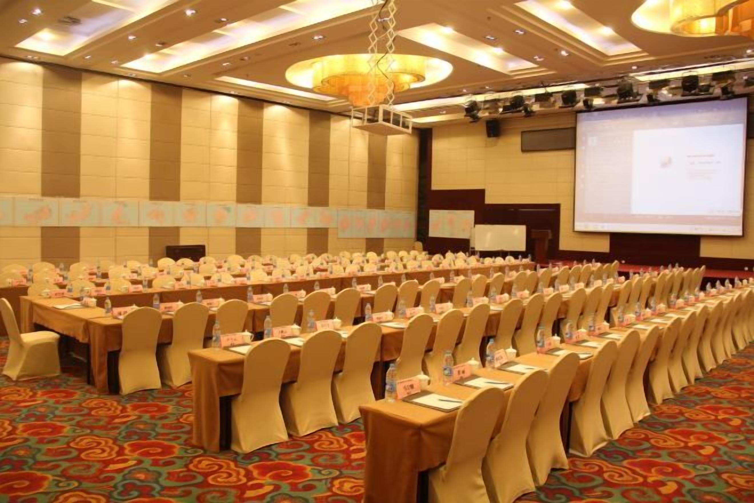 A conference room with rows of tables and chairs, a projector screen, and a carpeted floor.