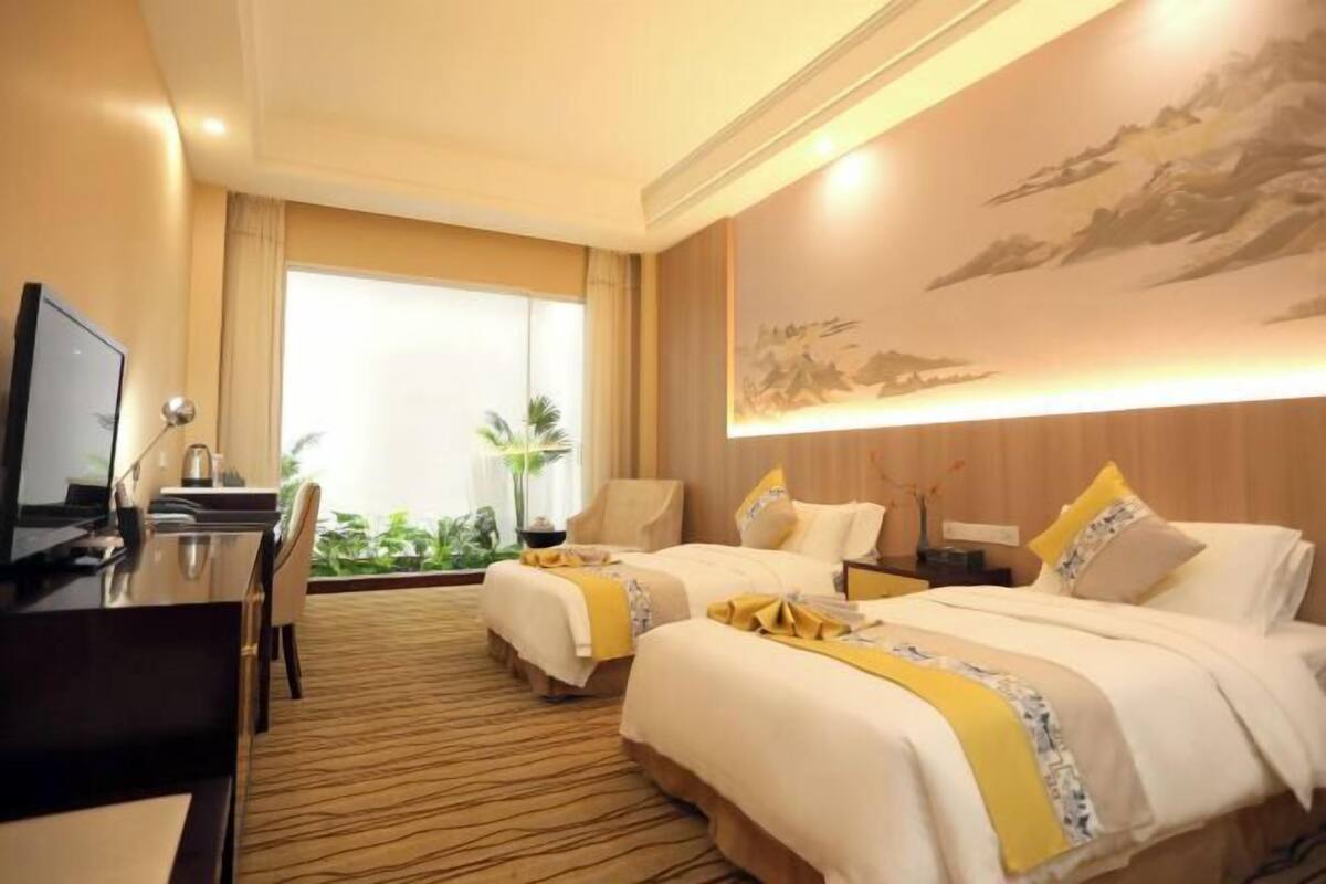 A hotel room with two beds, a desk, a TV, and a large window with a view of greenery.
