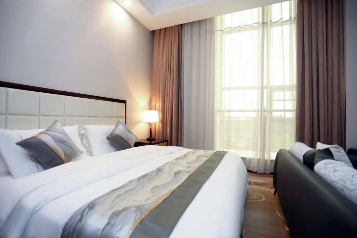 A hotel room with a large bed, a bedside table with a lamp, a sofa, and a window with curtains.