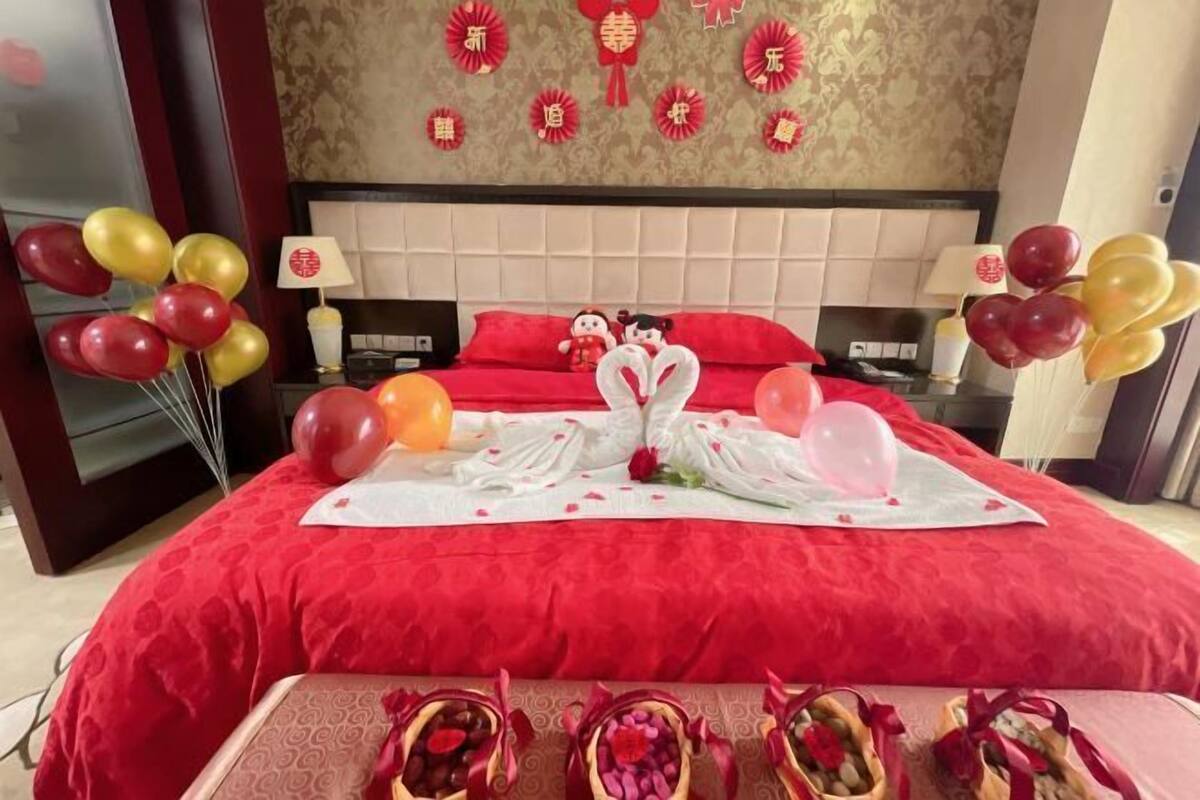 A hotel room with a red bedspread, decorative wall with Chinese characters, and a table with fruit baskets.