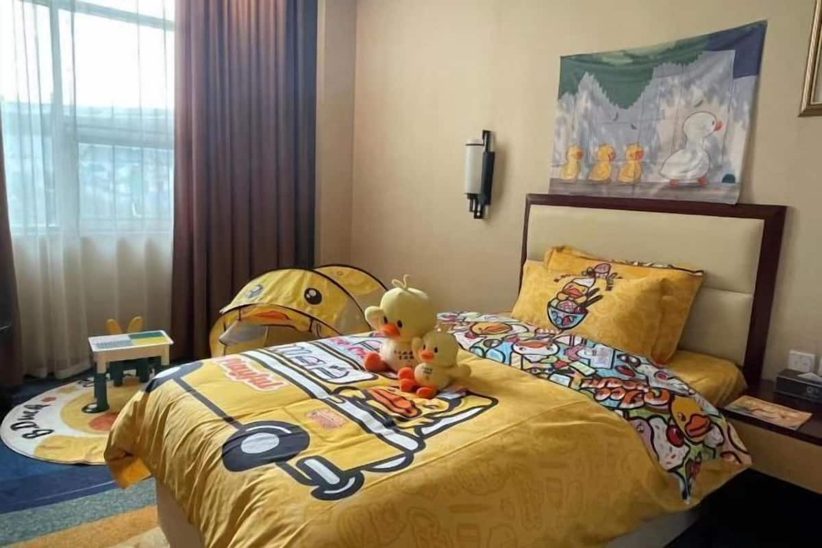 A hotel room with a bed featuring a cartoon-themed duvet, a wall-mounted light, a painting of ducks, and a small table with a toy.