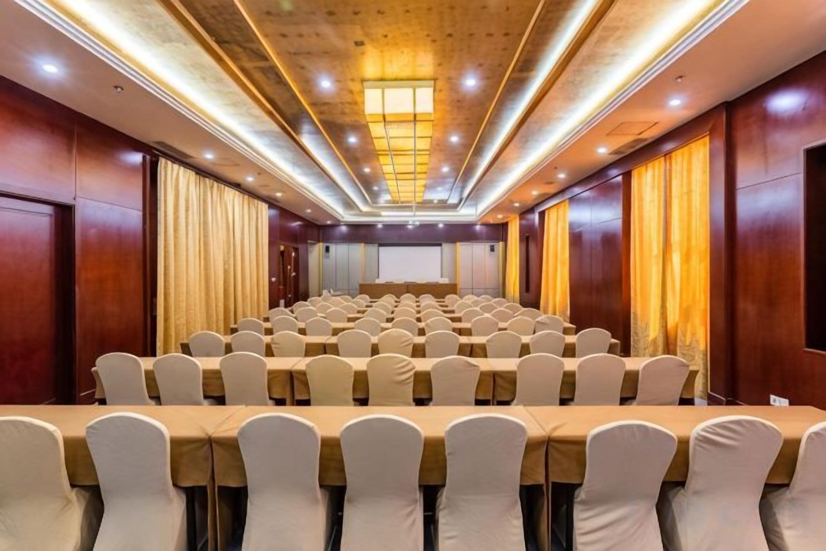 Un salón de conferencias con filas de mesas y sillas, un escenario al frente y paneles de madera en las paredes.